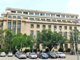 Facade of a large beige office building with rows of windows, green trees, and a parking lot in the foreground.