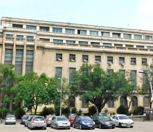Facade of a large beige office building with rows of windows, green trees, and a parking lot in the foreground.