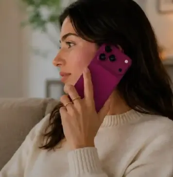 Woman sitting on a sofa, talking on a pink smartphone held to her ear, in a cozy living room with warm lighting.