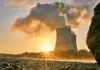 Industrial cooling towers releasing steam at sunset, seen from a rocky shoreline