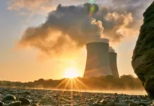 Industrial cooling towers releasing steam at sunset, seen from a rocky shoreline