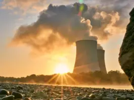 Industrial cooling towers releasing steam at sunset, seen from a rocky shoreline