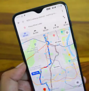 Google Maps Updates Aduc Multiple Schimbari cu AI de la Google Hand holds a smartphone showing a navigation app with colored routes and travel times on a city map