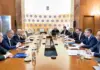 Formal boardroom meeting with executives seated along a long wooden table; microphones, documents and water glasses on the table.
