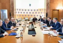 Formal boardroom meeting with executives seated along a long wooden table; microphones, documents and water glasses on the table.