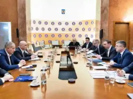 Formal boardroom meeting with executives seated along a long wooden table; microphones, documents and water glasses on the table.