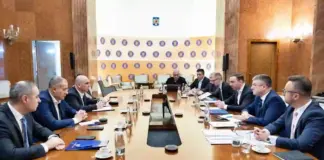 Formal boardroom meeting with executives seated along a long wooden table; microphones, documents and water glasses on the table.