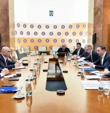 Guvernul Interimar Bolojan a Avertizat Oficial Comisia Europeana Formal boardroom meeting with executives seated along a long wooden table; microphones, documents and water glasses on the table.