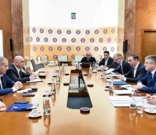 Formal boardroom meeting with executives seated along a long wooden table; microphones, documents and water glasses on the table.