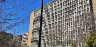 Tall rectangular high-rise office building with a grid of windows and visible window units, bare trees in foreground and a clear blue sky.