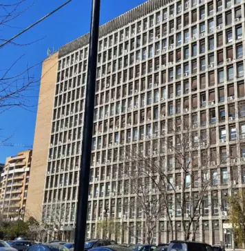 Tall rectangular high-rise office building with a grid of windows and visible window units, bare trees in foreground and a clear blue sky.