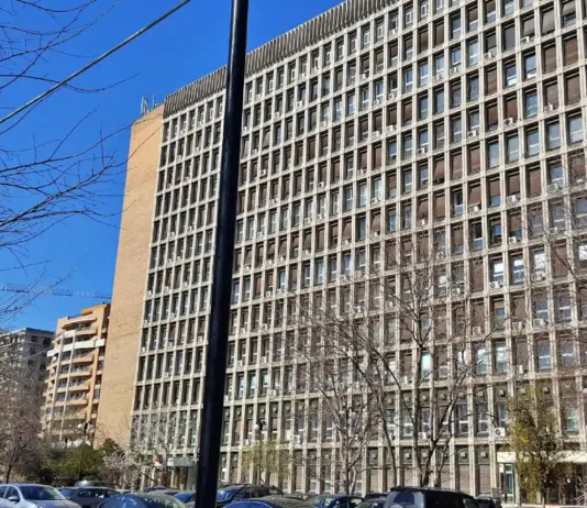 Tall rectangular high-rise office building with a grid of windows and visible window units, bare trees in foreground and a clear blue sky.