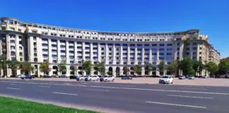 Curved white multi-story building with many windows and arches along a busy street, cars parked in front on a sunny day.