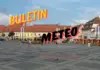 Prognoza Meteo ANM cu Estimarile Starii Vremii in Romania pentru 30 de Zile Town square with pastel buildings and people walking, overlaid with bold yellow and pink text reading 'BULETON METEO' across the sky.