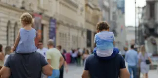 Two kids on an adult's shoulders walk through a crowded city street seen from behind.