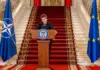 Romania Pregateste Trecerea la Moneda EURO, Apelul Lansat de Nicusor Dan Romanian president delivers a formal speech at a podium, flanked by Romanian, NATO, and EU flags on a grand staircase.
