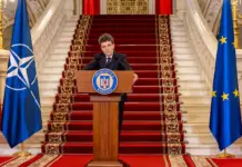 Romania Pregateste Trecerea la Moneda EURO, Apelul Lansat de Nicusor Dan Romanian president delivers a formal speech at a podium, flanked by Romanian, NATO, and EU flags on a grand staircase.