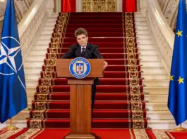 Romanian president delivers a formal speech at a podium, flanked by Romanian, NATO, and EU flags on a grand staircase.