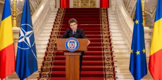 Romanian president delivers a formal speech at a podium, flanked by Romanian, NATO, and EU flags on a grand staircase.