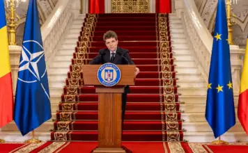Romanian president delivers a formal speech at a podium, flanked by Romanian, NATO, and EU flags on a grand staircase.