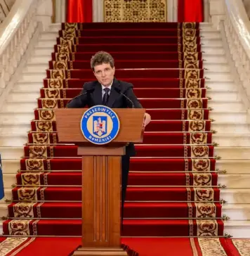 Romanian president delivers a formal speech at a podium, flanked by Romanian, NATO, and EU flags on a grand staircase.