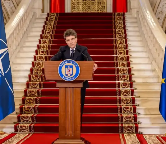 Romanian president delivers a formal speech at a podium, flanked by Romanian, NATO, and EU flags on a grand staircase.