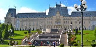 Grand historic palace with symmetrical towers, central staircase, and manicured gardens under a blue sky; visitors on the lawn.