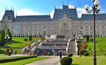 Grand historic palace with symmetrical towers, central staircase, and manicured gardens under a blue sky; visitors on the lawn.