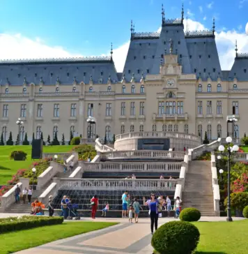 Starea de Alerta va Lasa in Bezna Unele Orase din Romania, Explicatiile Primarului din Iasi Grand historic palace with symmetrical towers, central staircase, and manicured gardens under a blue sky; visitors on the lawn.