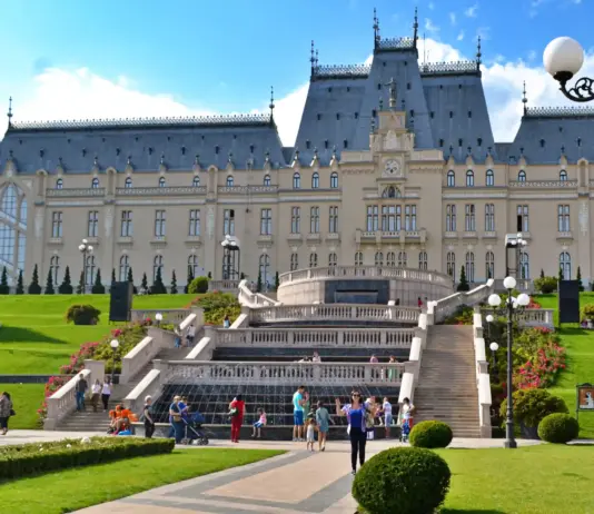 Grand historic palace with symmetrical towers, central staircase, and manicured gardens under a blue sky; visitors on the lawn.