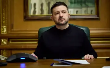 Volodimir Zelenski Trage un Semnal de Alarma: Zeci de Mii de Nord-Coreeni Asteptati in Rusia Man in a black shirt sits at a wooden desk in a formal office, looking off to the side with papers and a computer mouse in view.