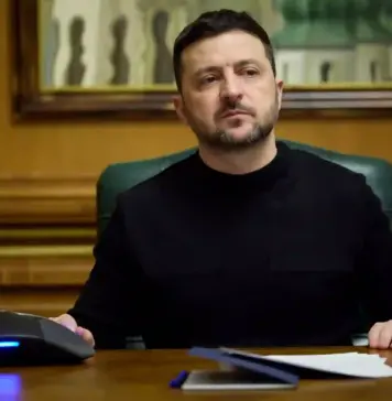 Volodimir Zelenski Trage un Semnal de Alarma: Zeci de Mii de Nord-Coreeni Asteptati in Rusia Man in a black shirt sits at a wooden desk in a formal office, looking off to the side with papers and a computer mouse in view.