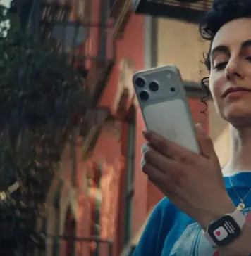 iOS 27 Confirma Aceste 6 Noi Modele de Telefoane iPhone Young woman outdoors, checking her smartphone, wearing a blue shirt and a crossbody bag.