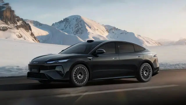Black sedan driving on a snow-covered mountain road with alpine peaks in the background.