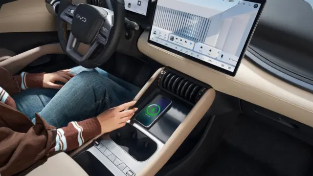 Person sits in a car with hands on a wireless charging pad in the center console while a large touchscreen display runs across the dashboard above the vents, showing car controls and media icons.