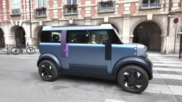 Blue futuristic car with a purple stripe drives along a city street in front of a brick building with arches and balconies.