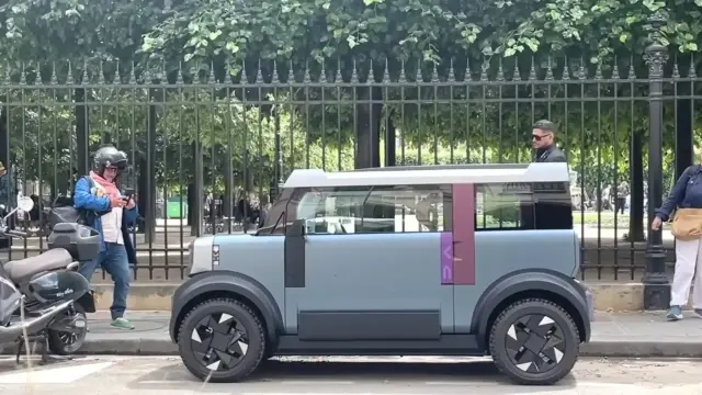 A futuristic gray compact vehicle with large black wheels parked along a curb, as a man wearing a helmet and blue jacket stands nearby and an older woman with a tan bag stands by a lamppost, with a tall black iron fence and green trees behind.
