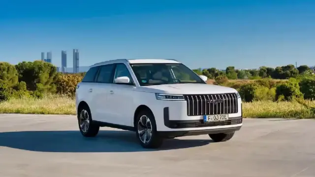 White SUV parked on a paved surface with green fields and a distant city skyline under a blue sky, bright and clear.