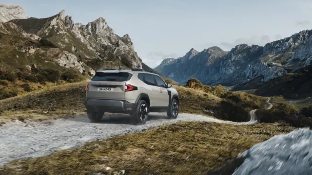 SUV driving along a dirt road through a rugged alpine landscape with jagged mountains behind it.